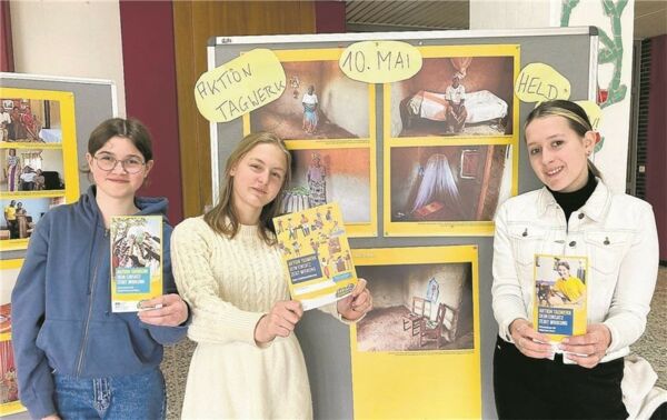 3 Schülerinnen präsentieren den Informationsflyer zur "Aktion Tagwerk" vor der Wanderausstellung zum Thema "Leben in Ruanda".