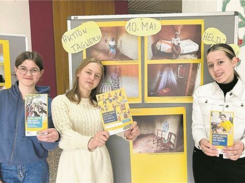 3 Schülerinnen präsentieren den Informationsflyer zur "Aktion Tagwerk" vor der Wanderausstellung zum Thema "Leben in Ruanda".